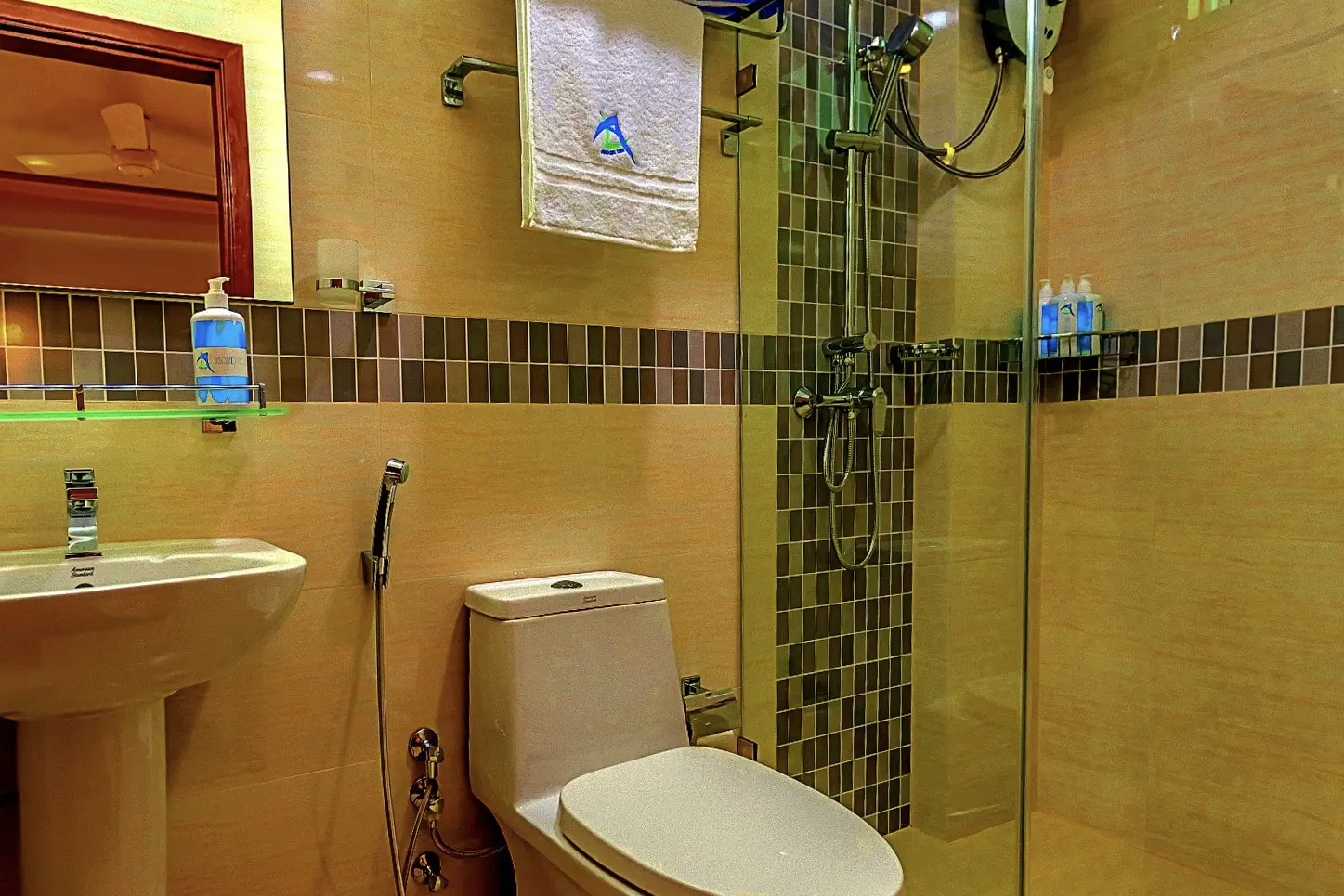 A modern hotel bathroom features beige tile, a pedestal sink, and a shower with mosaic accents.